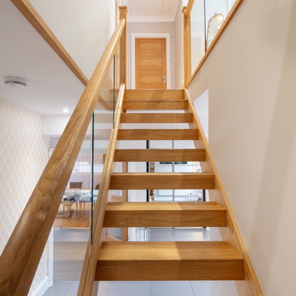 Looking up an oak staircase that has open risers and embedded glass balustrades. An oak door is just visible on the landing at the top of the flight.